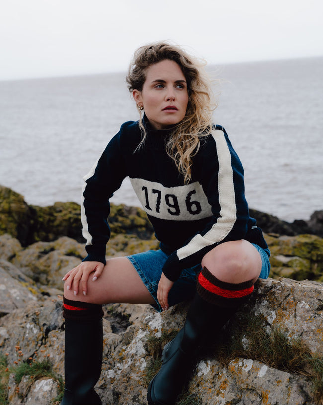 Woman sitting on rocks by the sea wearing a black sweater with '1796' print.