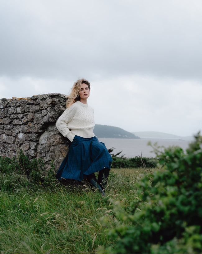 Woman in a white sweater and blue skirt leaning against a stone wall in a scenic landscape.