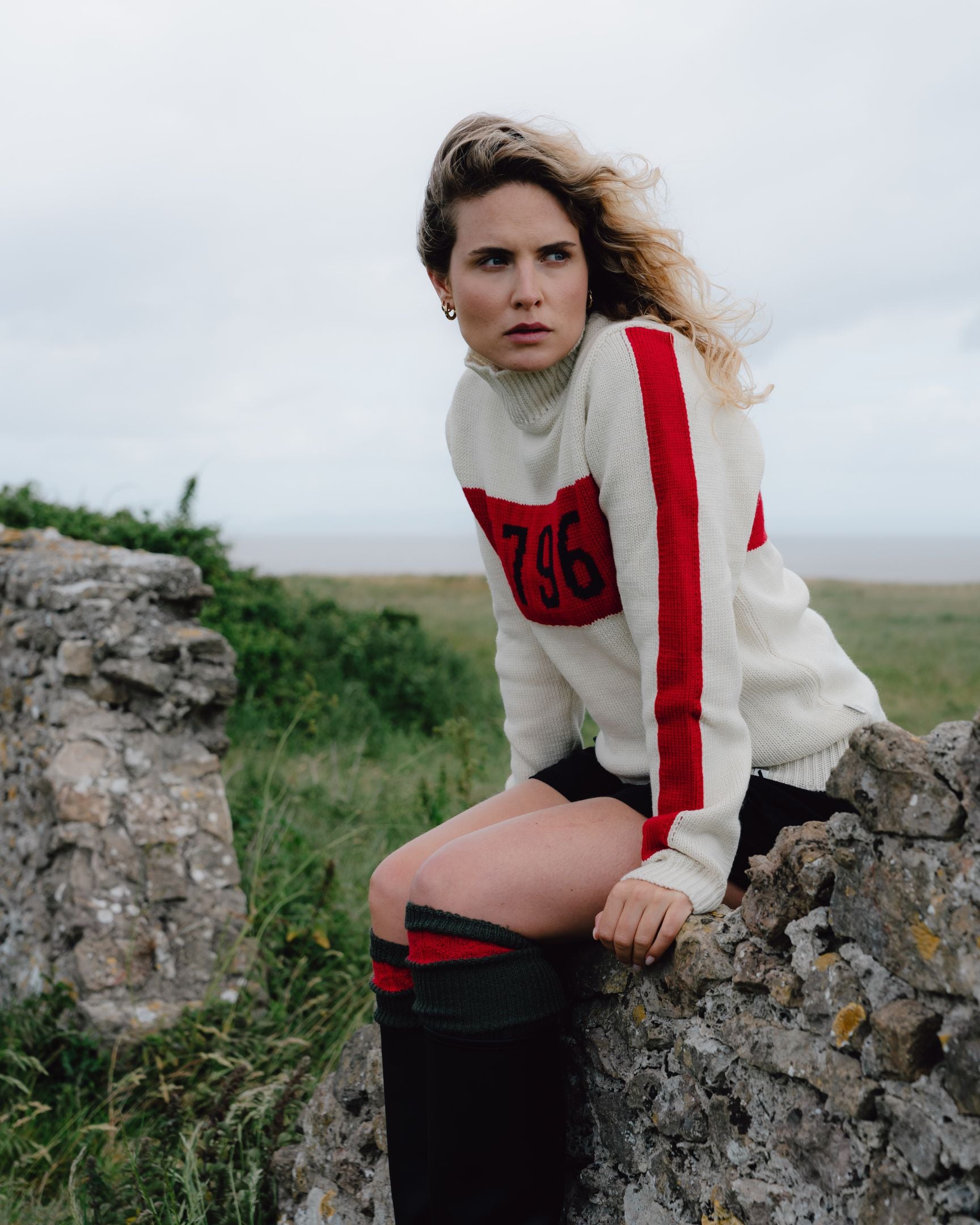 Woman wearing a white sweater with red and black details, sitting on rocks in a natural setting.
