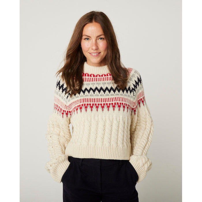 Flatlay image of a cream cable knitted, full length sleeve and crew neck jumper. The top half of the jumper has red, light grey and navy horizontal nordic knit patterns.