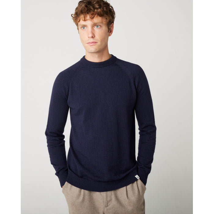 A flat lay image of a navy blue crew neck jumper. The jumper has a fine knit with a ribbed crew neck and hem and has full length sleeves. A male model wearing the navy jumper with a pair of beige trousers and black shoes.  The back of the model wearing the same outfit.