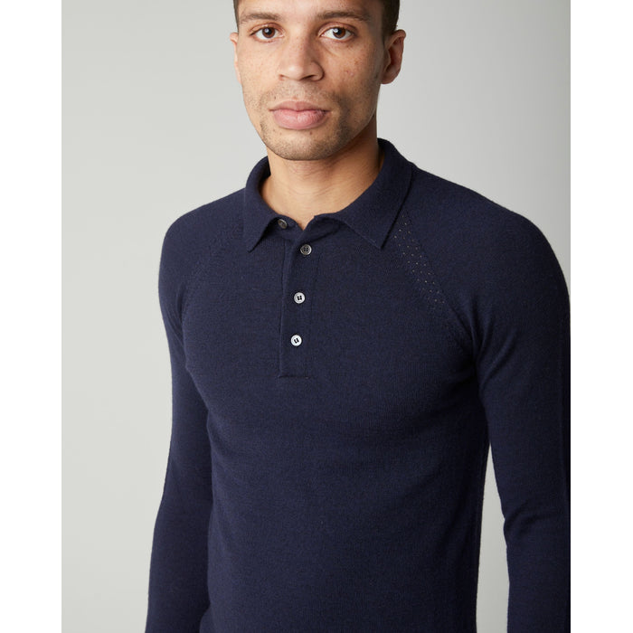A flatlay image of a navy blue polo shirt jumper. The jumper has a collar and  4 buttons to the chest. The hem and cuff have a ribbed detail. The jumper is a light weight, fine knit jumper and only one colour all over. A male model wearing the jumper and looking of to the left. A male model wearing the jumper outside whilst walking across a wooden beam in a field. He is wearing the jumper with beige trousers and black shoes.