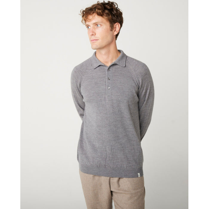 A flatlay image of a grey polo shirt jumper. The jumper has a collar and  4 buttons to the chest. The hem and cuff have a ribbed detail. The jumper is a light weight, fine knit jumper and only one colour all over. A male model wearing the jumper with all of the button done up and looking to the right.