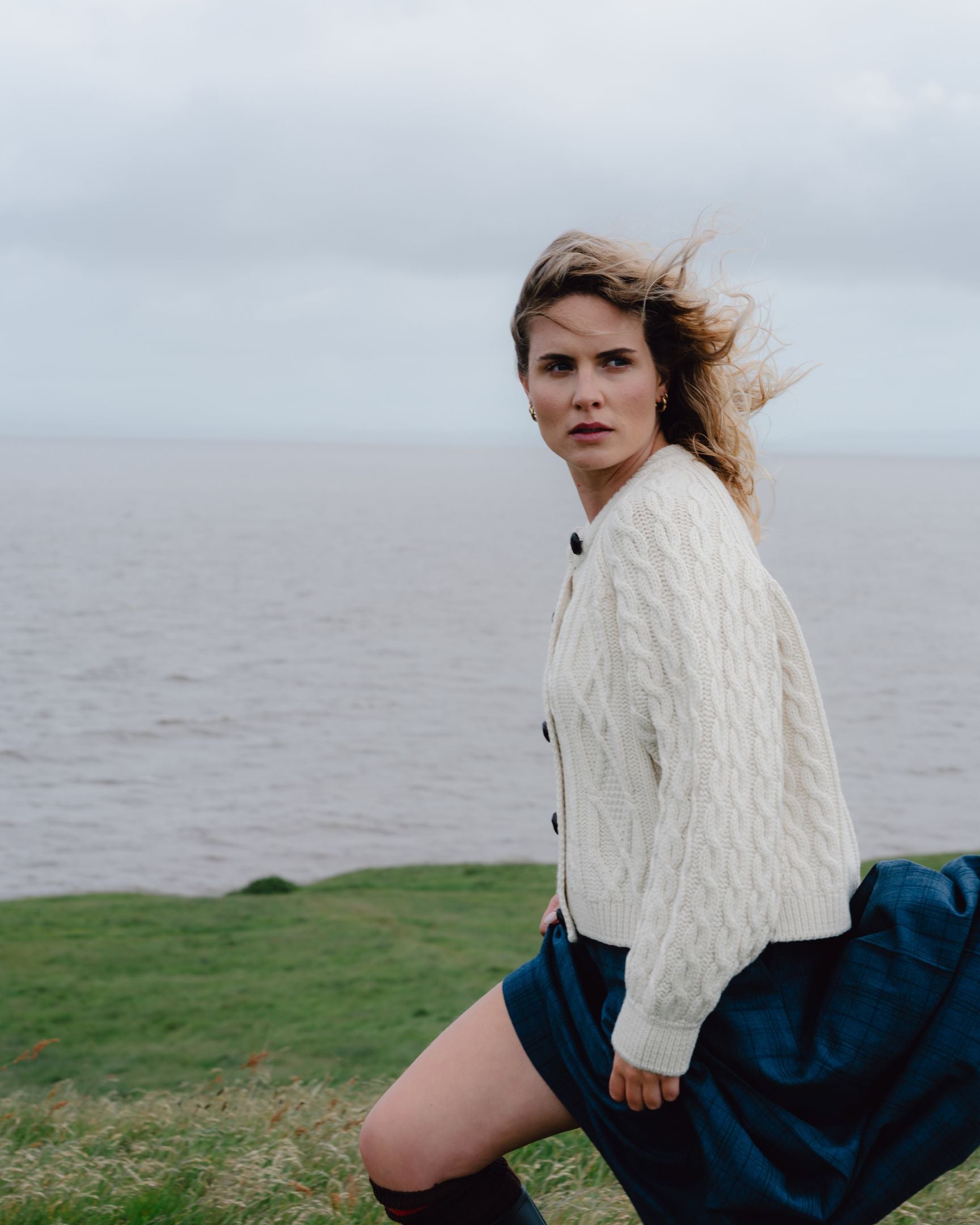 Woman in a white cable knit sweater and blue skirt standing by a body of water.