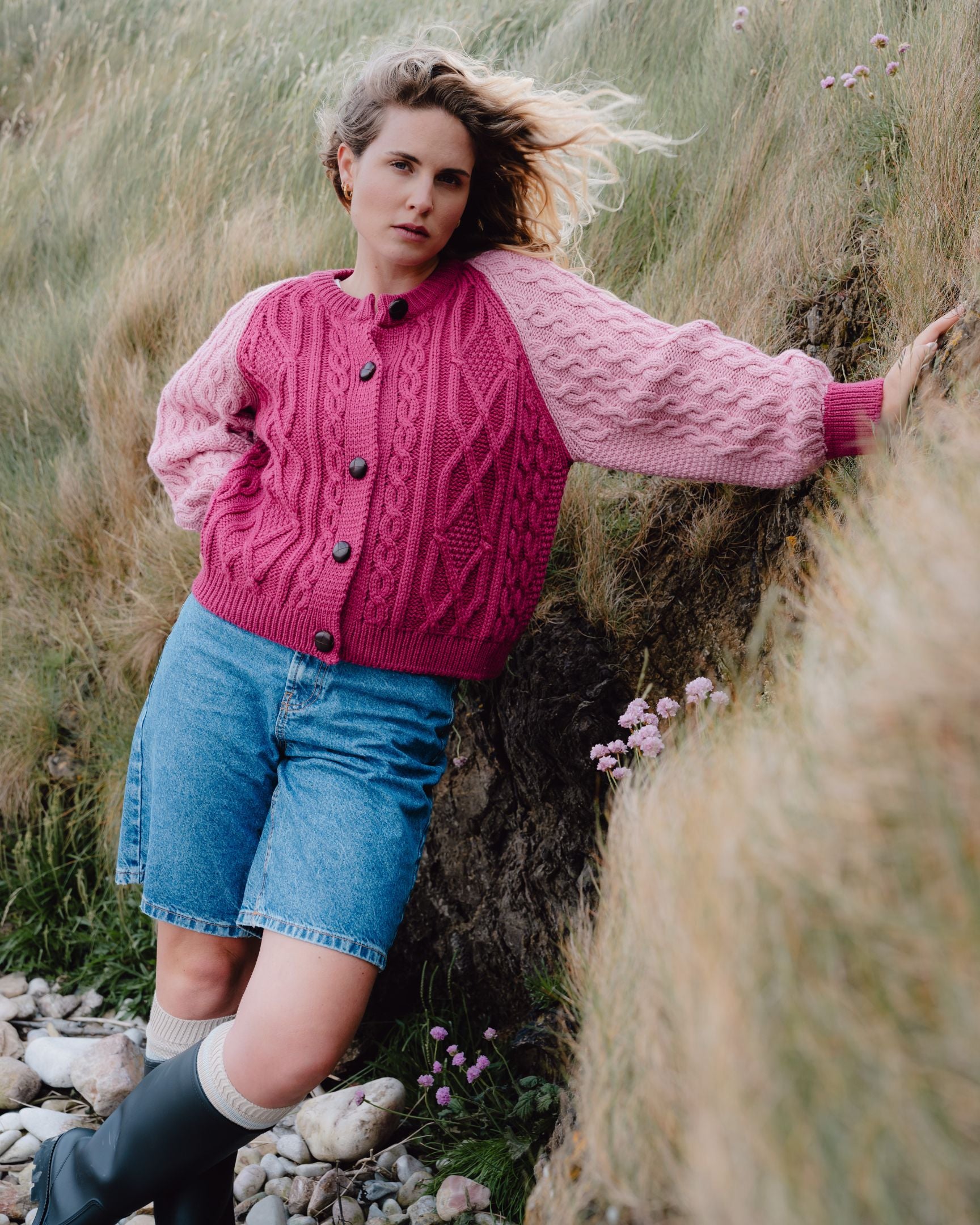 Woman wearing a pink cardigan and denim shorts standing in a natural setting with grass and rocks.