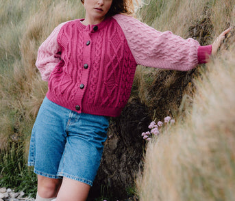 Woman wearing a pink cardigan and denim shorts standing in a natural setting with grass and rocks.