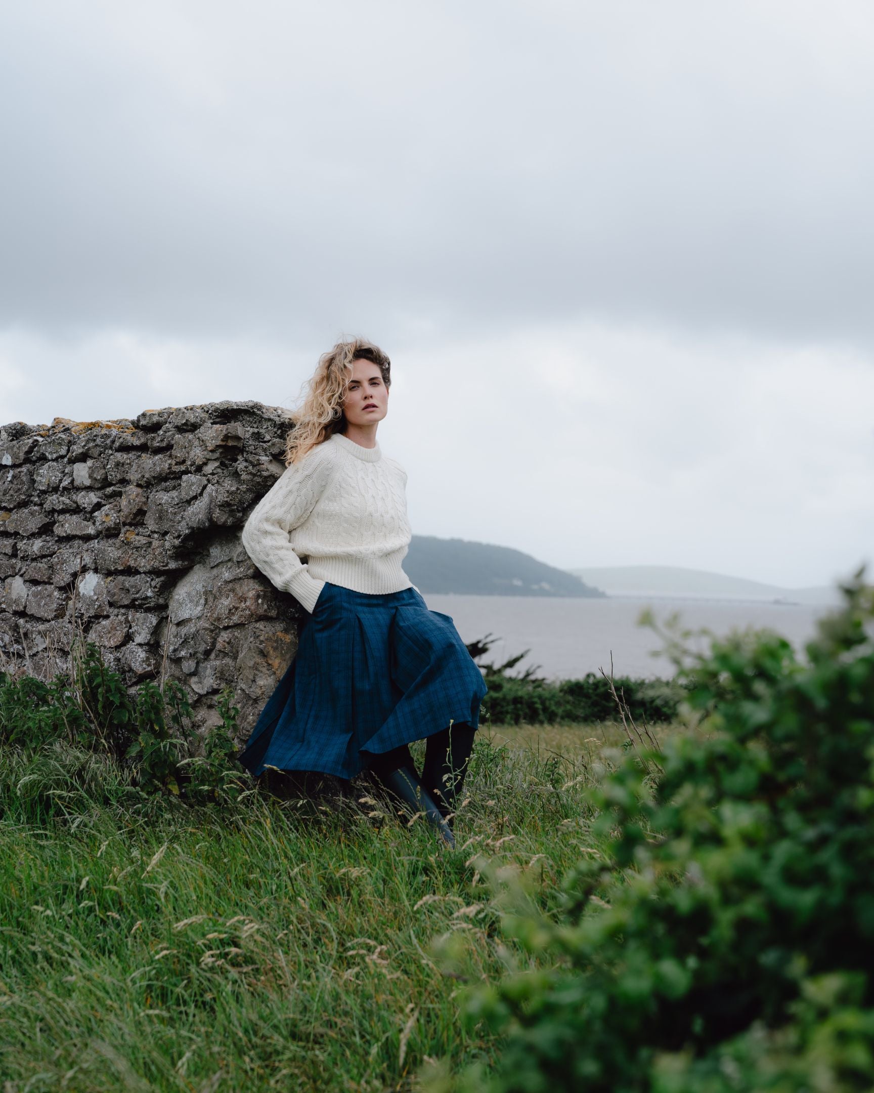 Woman in a white sweater and blue skirt leaning against a stone wall in a scenic landscape.