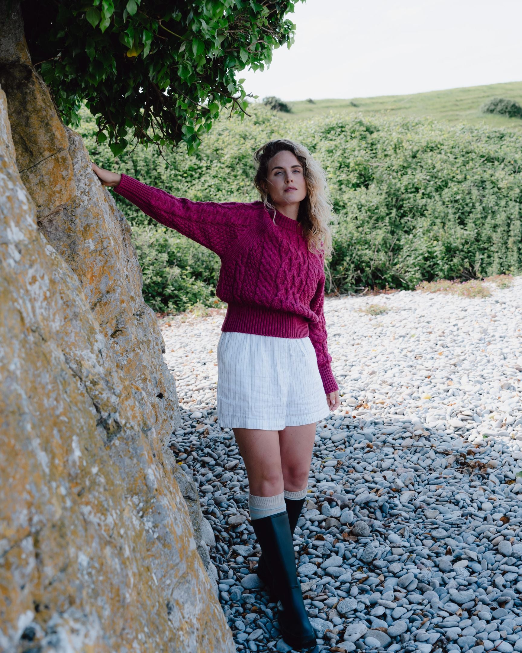 Woman in a pink sweater and white shorts standing near a stone wall with greenery in the background
