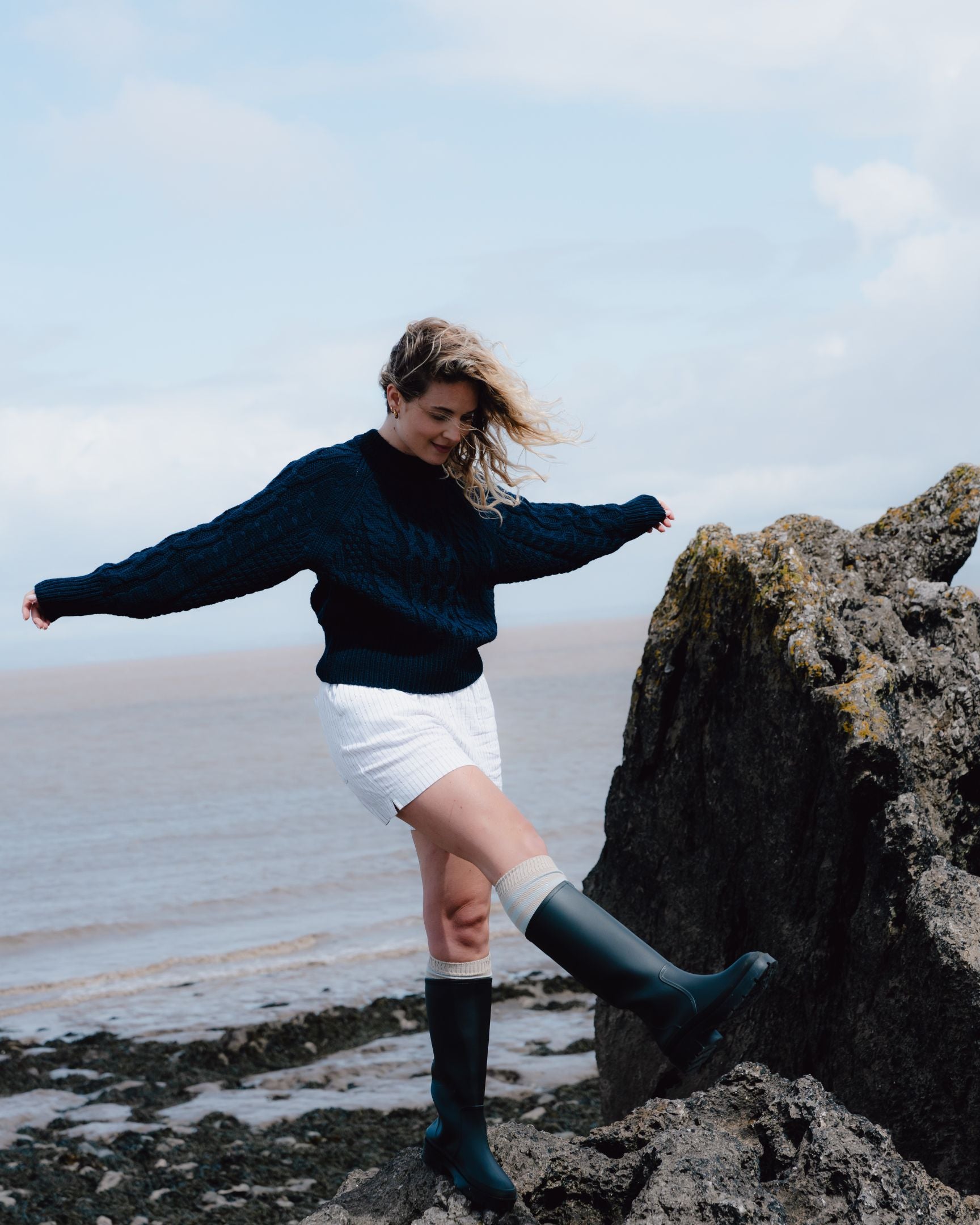 Person standing on a rock by the water wearing a navy sweater, white shorts, and dark rain boots.