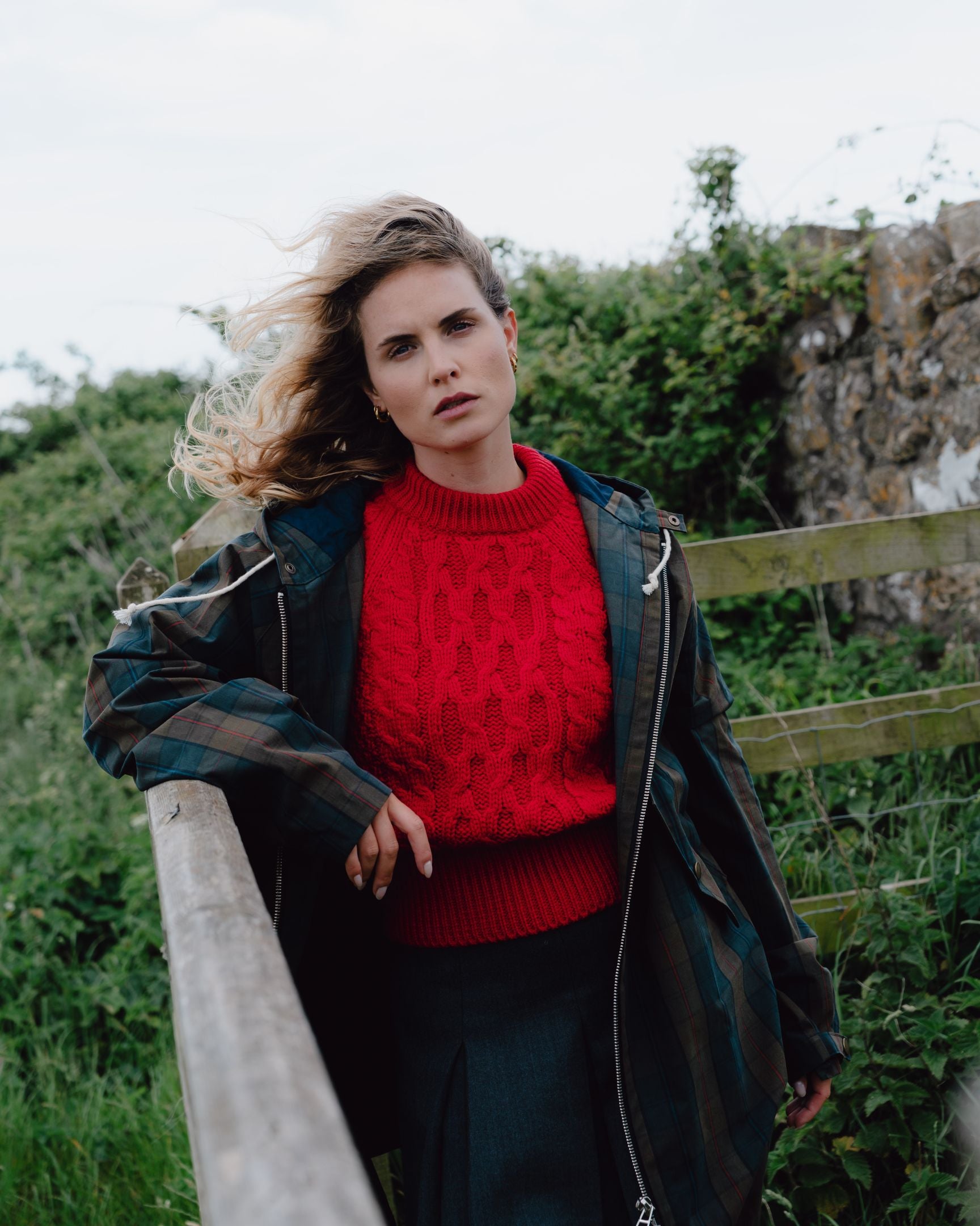 Woman wearing a red sweater and dark coat standing outdoors with greenery in the background