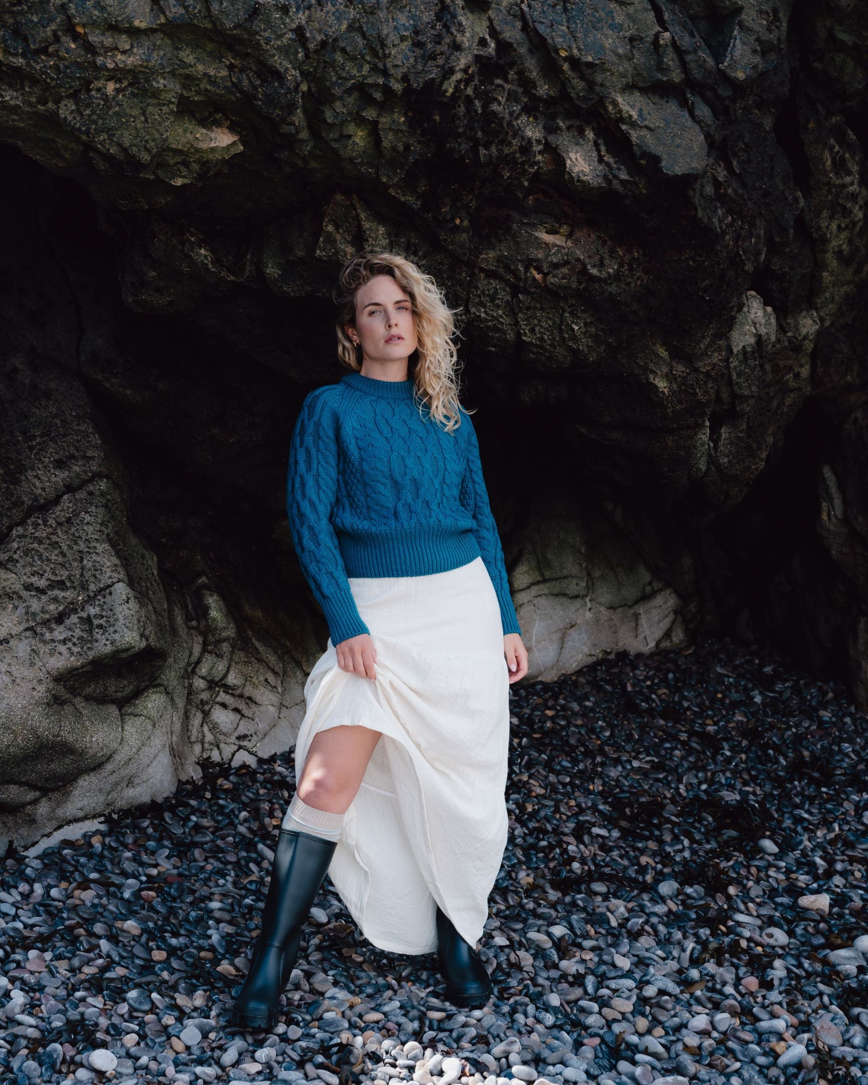 Woman in a blue sweater and white skirt standing in a rocky cave.