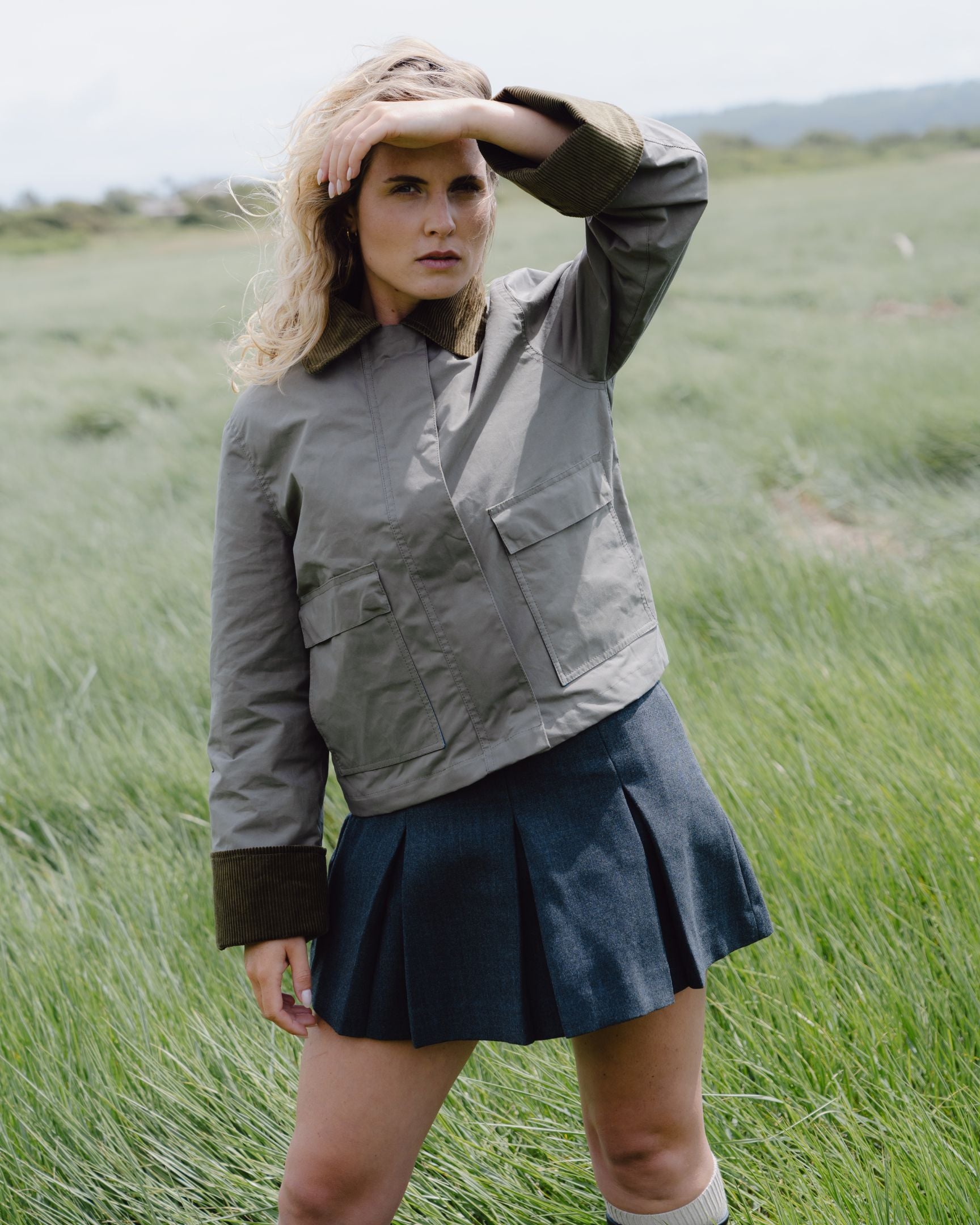 Woman wearing a green jacket and navy skirt standing in a grassy field.