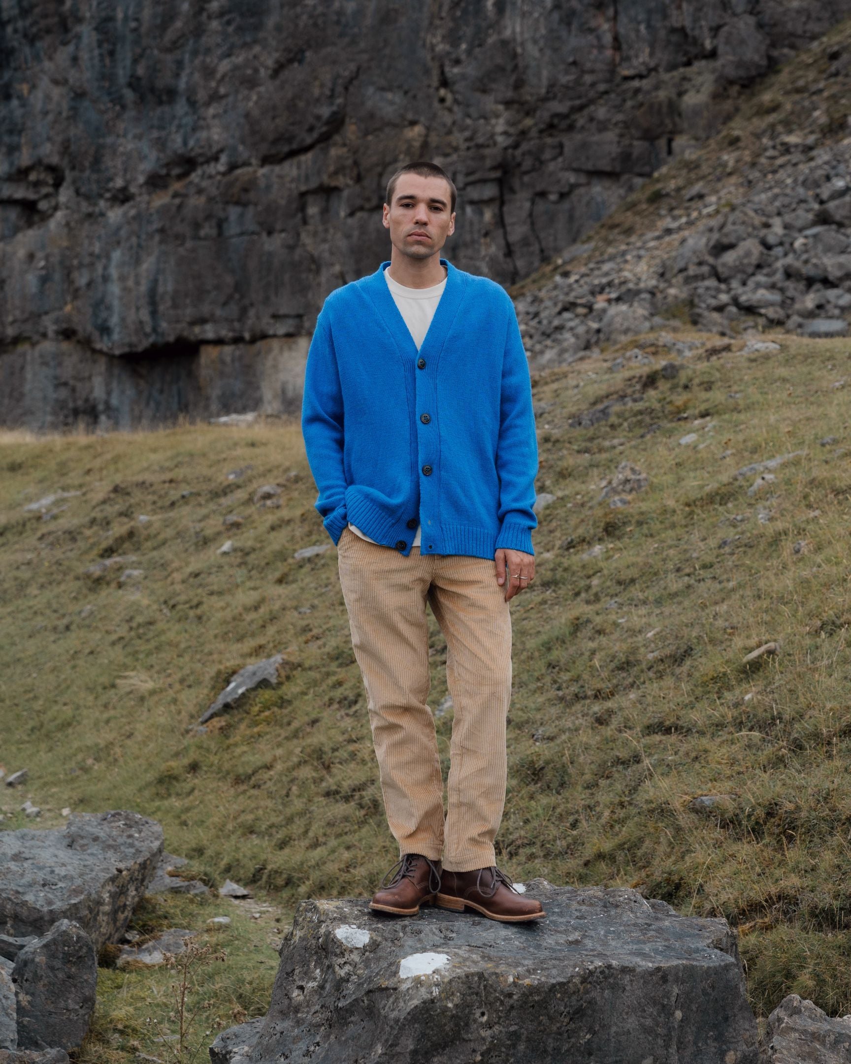 Man wearing a blue cardigan and beige pants standing on a rocky outcrop with a mountainous background.