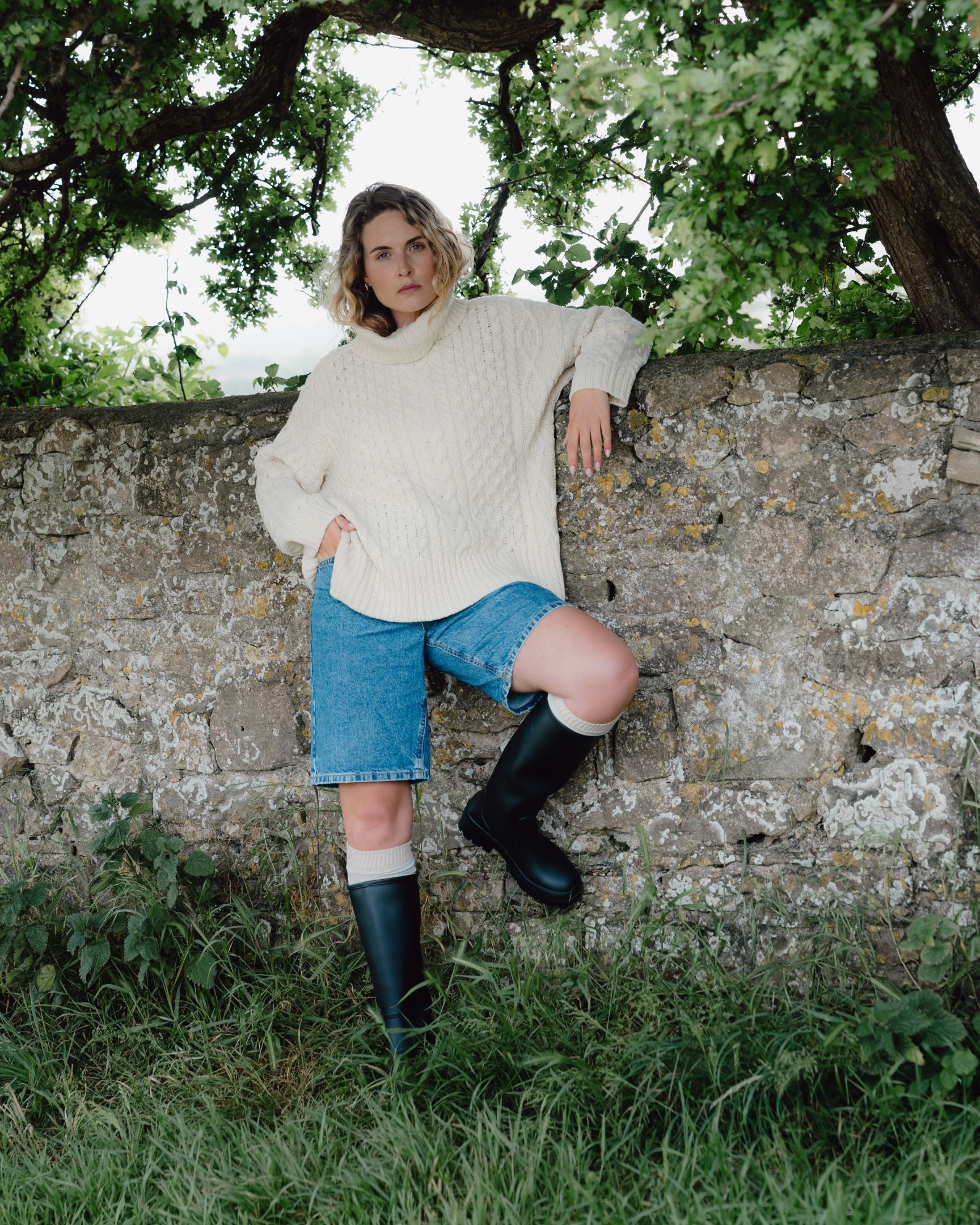 Woman in a white sweater and blue shorts standing against a stone wall with greenery around.