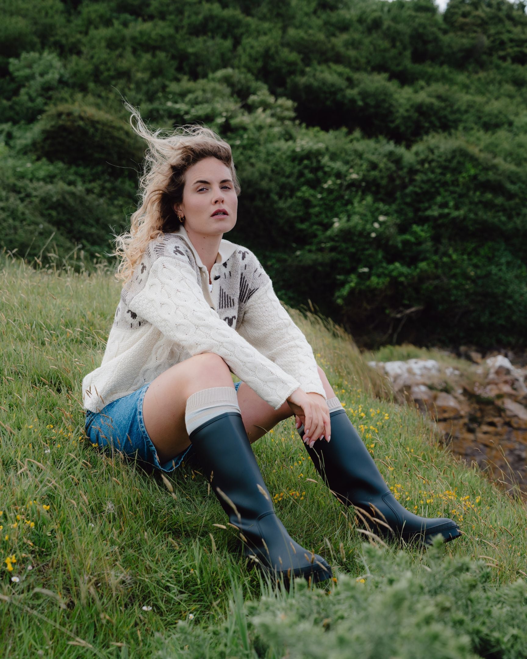 Woman sitting on a grassy hill wearing a white shirt, denim shorts, and green rain boots.