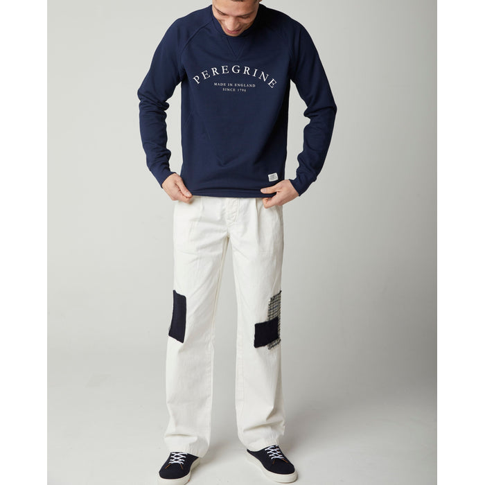 Flatlay image of cream straight leg trousers with a black button and belt loops on the waistband, the trousers have a small navy horizontal patch of wool sewn onto the right knee and on the left knee there is a smaller vertical navy patch of wool sewn overlaying a wool patch of navy, black, grey and white dog tooth print.